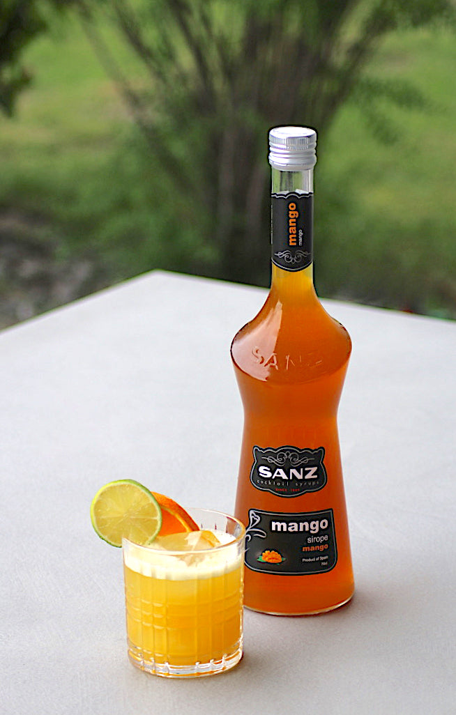 Flasche Sanz Mangosirup neben Tropical Fling Cocktail mit Limetten- und Orangenscheibe, serviert auf Eis vor einem unscharfen, grünen Hintergrund.