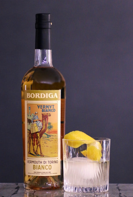 Flasche Bordiga Vermouth di Torino Bianco, 75 cl, mit Glas Vermouth auf Eis und Zitronenzeste, vor dunklem Hintergrund.