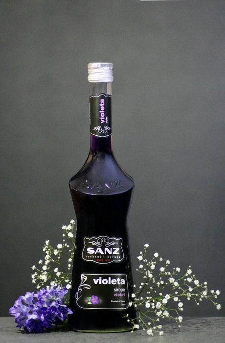 Flasche Sanz Violet-Sirup mit violettem Sirup, umgeben von violetten und weißen Blumen vor einem grauen Hintergrund.