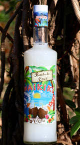 Flasche Batida de Coco Waikiki vor einem Baumstamm, mit tropischem Design und Etikett, das Kokosnüsse und Strandmotive zeigt.