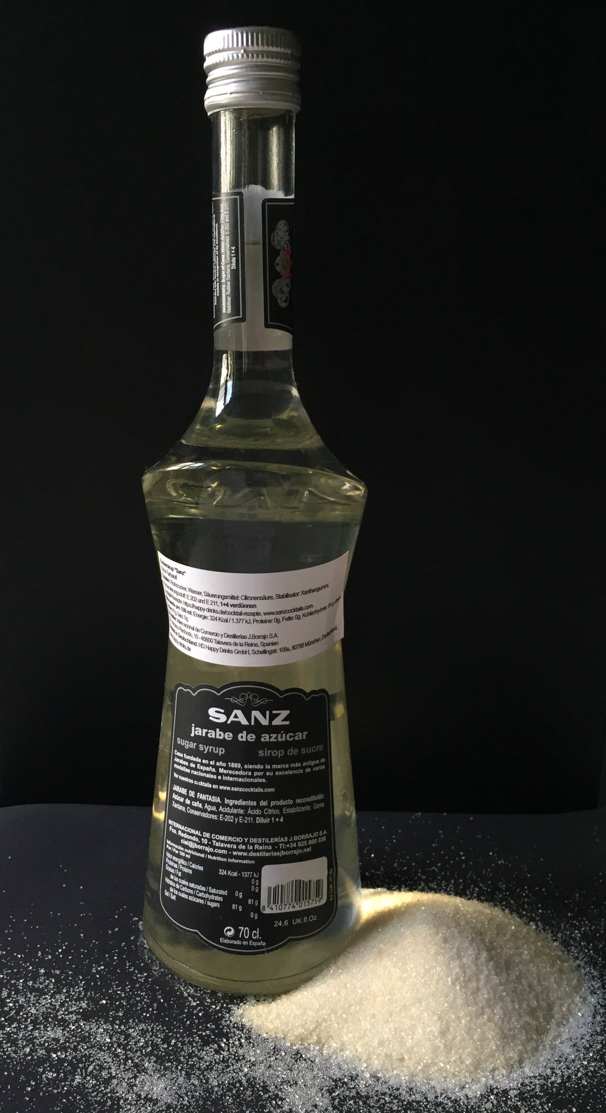 Rückseite der Flasche Sanz weißer Rohrzucker-Sirup, 70 cl, mit Angaben zu Inhaltsstoffen, auf schwarzem Hintergrund mit losem Zucker