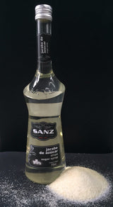 Flasche weißer Rohrzuckersirup von Sanz vor schwarzem Hintergrund, daneben ein Haufen kristalliner Zucker.